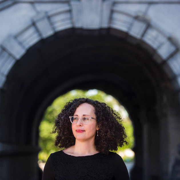 Victoria Van Hyning, a woman with light complexion, dark curly hair, and glasses standing in front of a stone arch. 